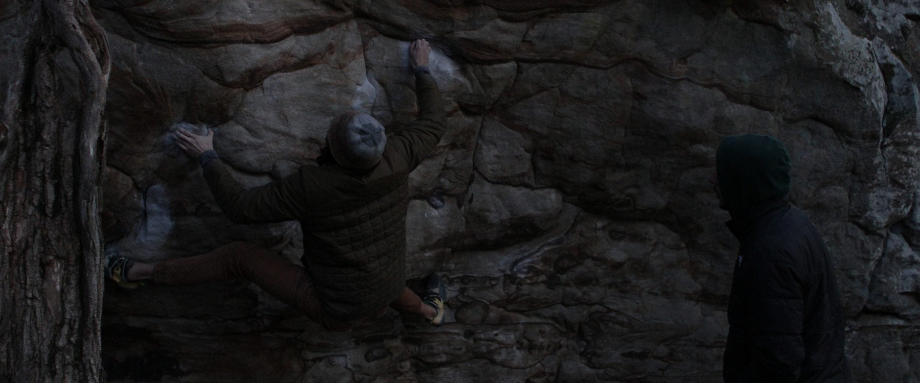 Climber on a sandstone boulder problem at dusk with a spotter watching from the leaf-covered landing