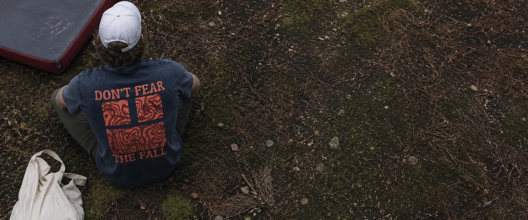 Climber resting on mossy ground in an Avila Climbing Co. Don't Fear The Fall tee beside a crash pad and tote bag