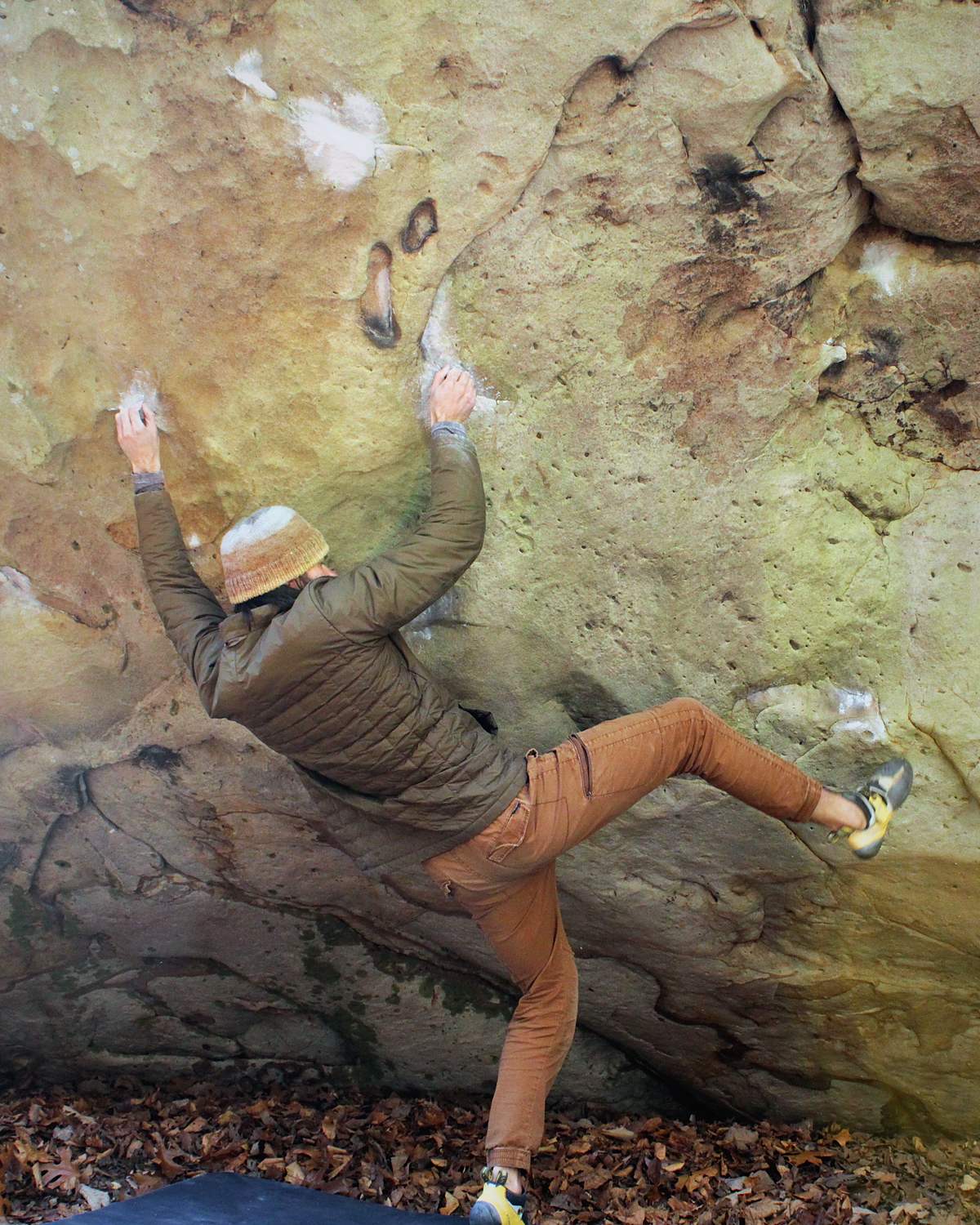 A person climbing a large rock wall, wearing a brown puffer jacket, brown pants, and climbing shoes, with a protective mat on the ground below.