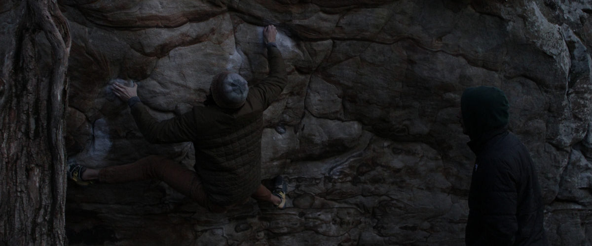 A person in a brown puffer jacket and beanie climbs a rocky outcrop, while another person in a dark hoodie watches from the right side of the frame.