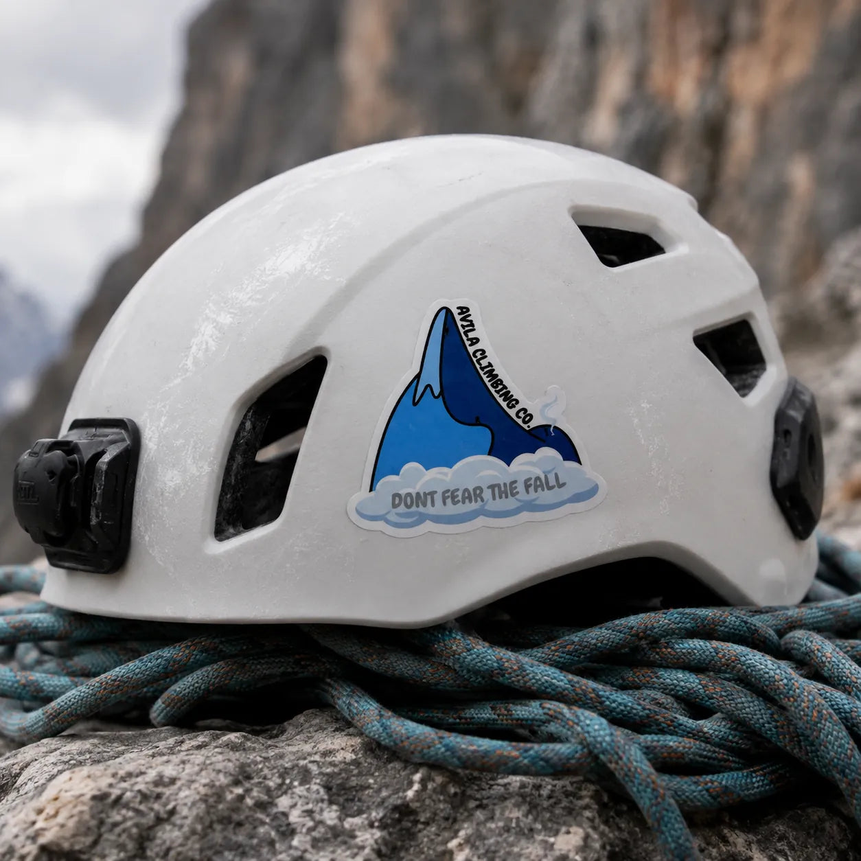 A white climbing helmet with a black mounting device on the side, resting on blue and gray climbing rope outdoors against a rocky mountain background. The helmet features a sticker with a blue mountain graphic and the text 'DON'T FEAR THE FALL' and 'Avila CLIMBING CO.