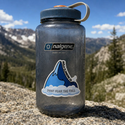 A translucent dark gray Nalgene water bottle with a blue lid and white carry loop, standing on a rock with a mountain landscape in the background. The bottle has a Nalgene logo sticker and a blue mountain sticker that reads 'Avila Climbing CO' and 'DONT FEAR THE FALL'.