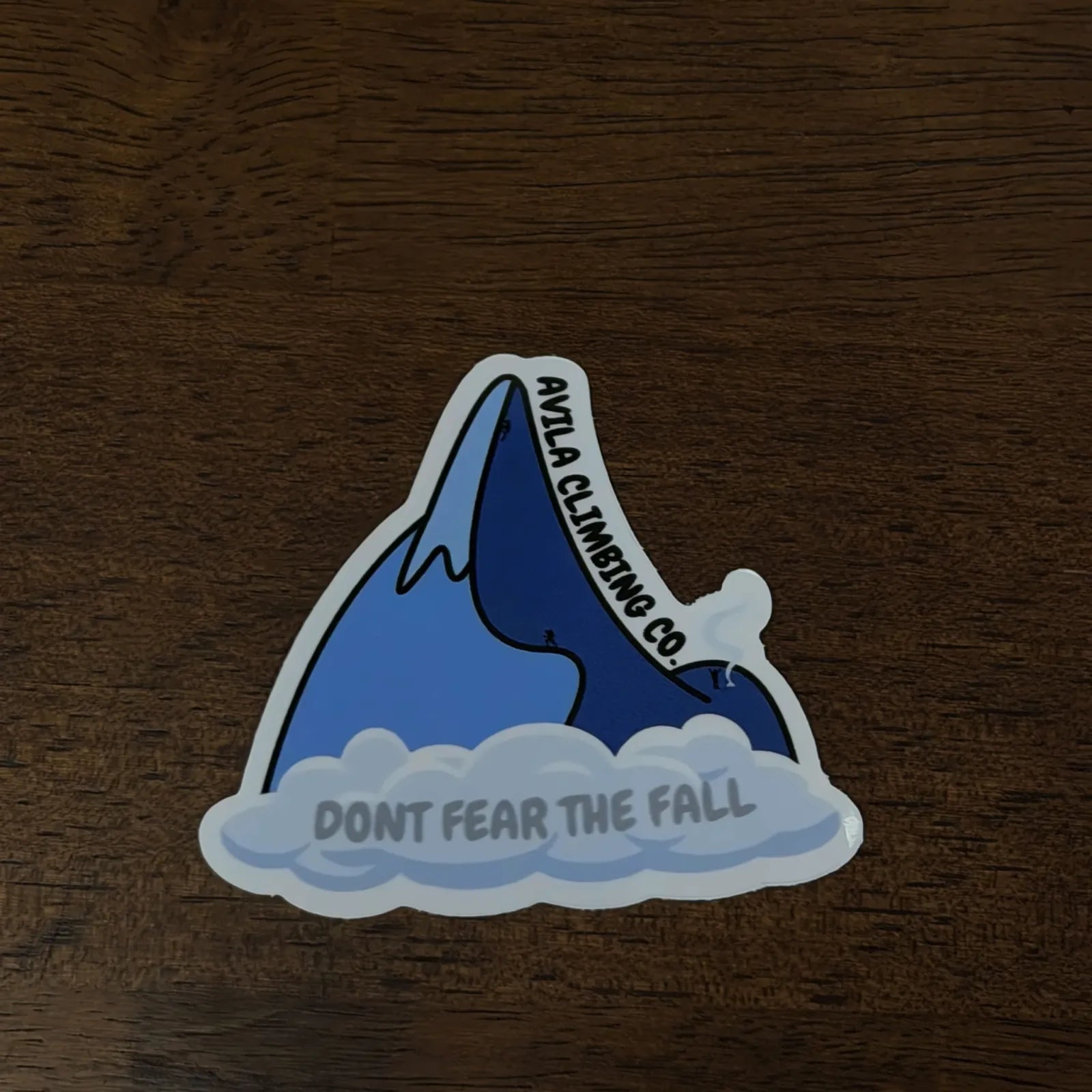 A die-cut vinyl sticker featuring a blue climbing peak above white clouds, with the text 'avilA CLIMBING CO.' on the mountain and 'DONT FEAR THE FALL' on the clouds, placed on a dark wood surface.