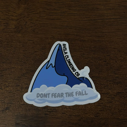 A die-cut vinyl sticker featuring a blue climbing peak above white clouds, with the text 'avilA CLIMBING CO.' on the mountain and 'DONT FEAR THE FALL' on the clouds, placed on a dark wood surface.